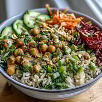A close-up of the Peanut Chickpea Rice Bowl shows fluffy brown rice topped with chickpeas, shredded carrots, and red cabbage, drizzled with creamy peanut dressing.